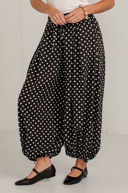 A person wearing loose black harem-style pants with white polka dots, paired with a white top and black flats, standing against a plain light gray wall.
