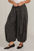 A person wearing loose black harem-style pants with white polka dots, paired with a white top and black flats, standing against a plain light gray wall.