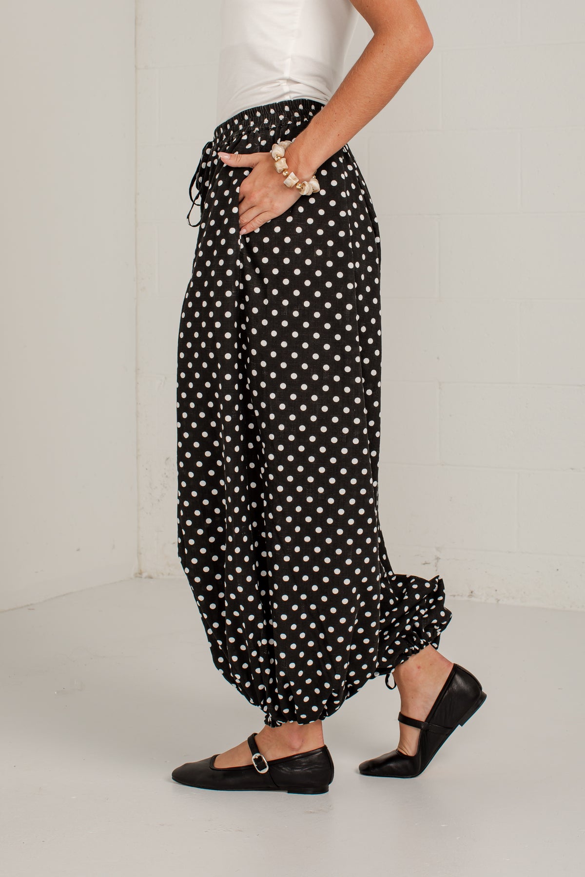 A person wearing loose black harem-style pants with a white polka dot pattern, paired with a white top and black Mary Jane shoes, standing against a plain light wall.