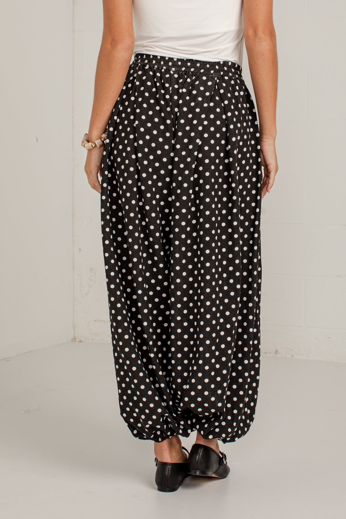 A person shown from the back wearing loose, balloon-style black pants with a white polka dot pattern, paired with a white top and black flats, in a plain indoor studio setting.