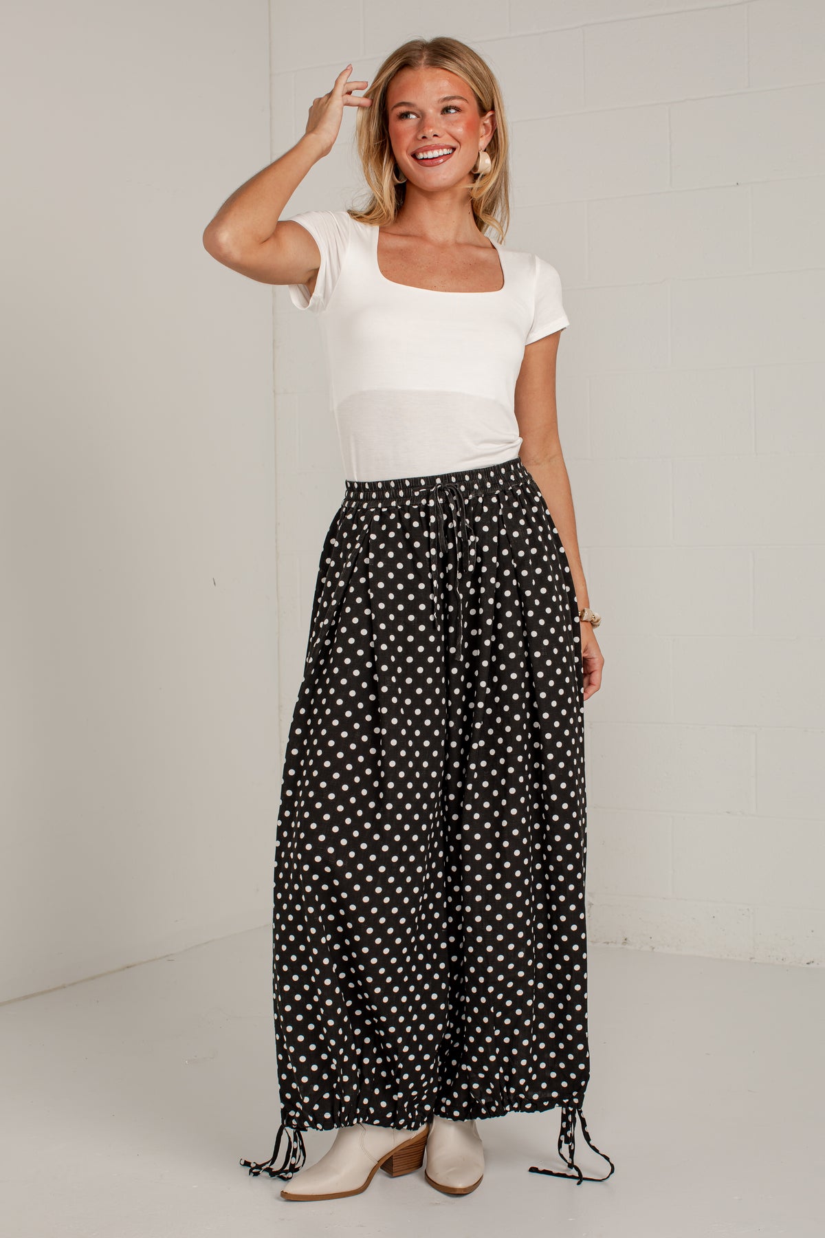 A woman modeling a white short-sleeve top and black wide-leg pants with a white polka dot pattern, standing against a white brick wall.