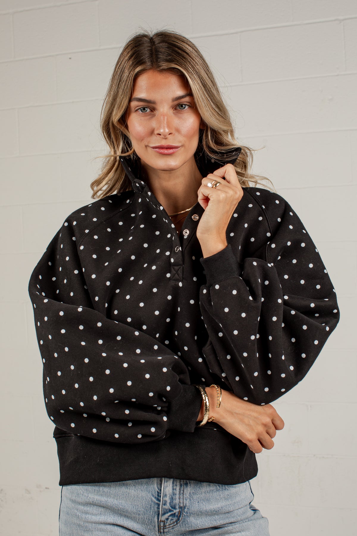 A woman wearing a black long-sleeve bomber-style jacket with white polka dots, paired with light blue jeans, standing against a white brick wall.