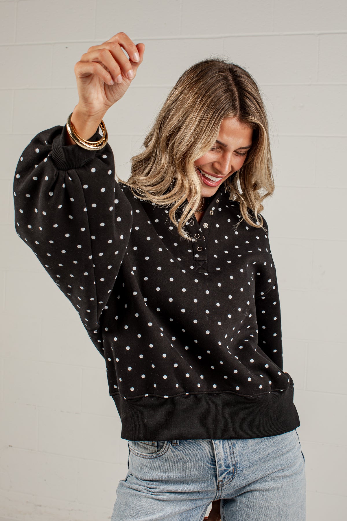 A woman wearing a black long-sleeve sweatshirt with white polka dots and light blue jeans, smiling and posing against a white brick wall.