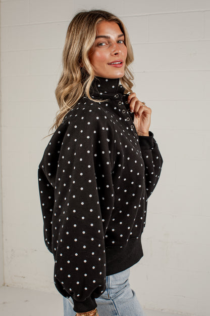A woman wearing a black turtleneck sweatshirt with white polka dots, paired with light blue jeans, standing against a white brick wall.