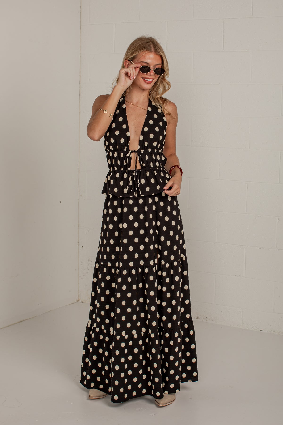 A woman modeling a black halter-neck maxi dress with a beige polka dot pattern, standing against a white brick wall.