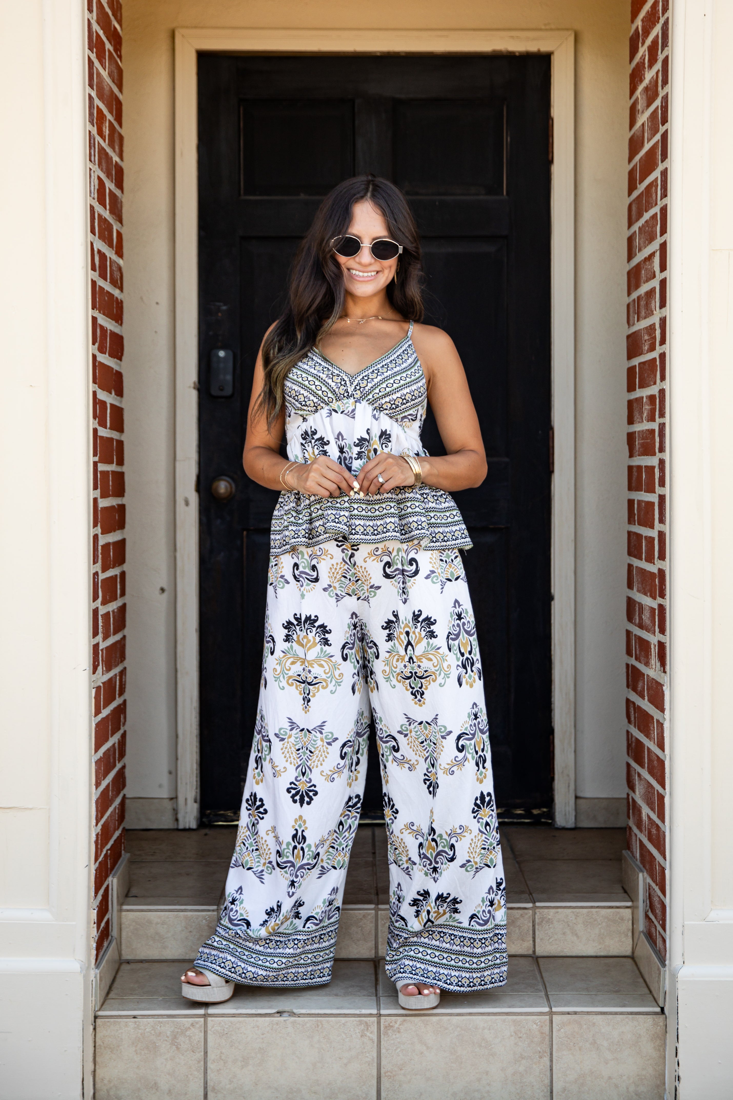 A woman wearing a sleeveless white jumpsuit with a navy and gold bohemian-style floral print stands on the tiled steps of a doorway framed by brick columns.