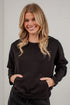 A smiling blonde woman wearing a black crewneck sweatshirt with a front kangaroo pocket, standing against a white brick wall.
