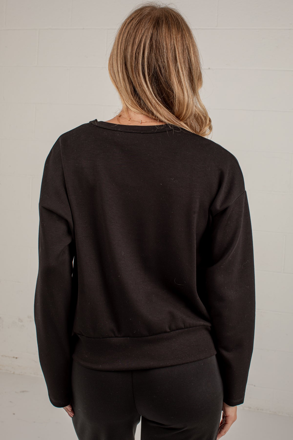 A woman viewed from behind wearing a dark brown crewneck sweatshirt and black pants against a white brick wall.