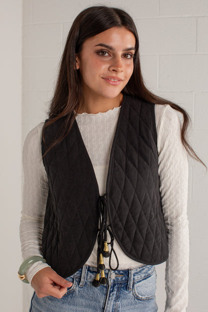 A woman wearing a black diamond-quilted sleeveless vest over a white ribbed long-sleeve top and blue jeans, standing against a white brick wall.