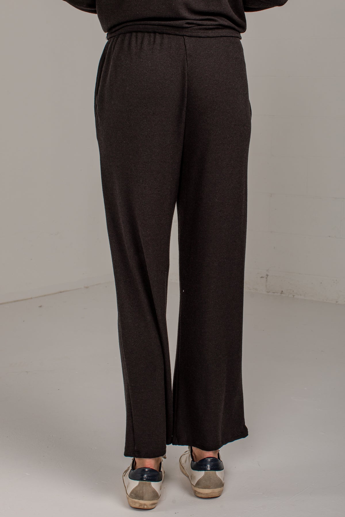 A person shown from the back wearing dark brown wide-leg knit pants and beige sneakers with navy accents, standing against a plain light gray studio background.