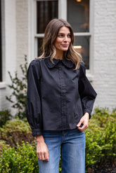 Woman wearing a black blouse with a ruffled collar and blue jeans standing outdoors.