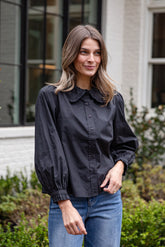 Woman wearing a black blouse with a high collar and blue jeans standing outdoors.