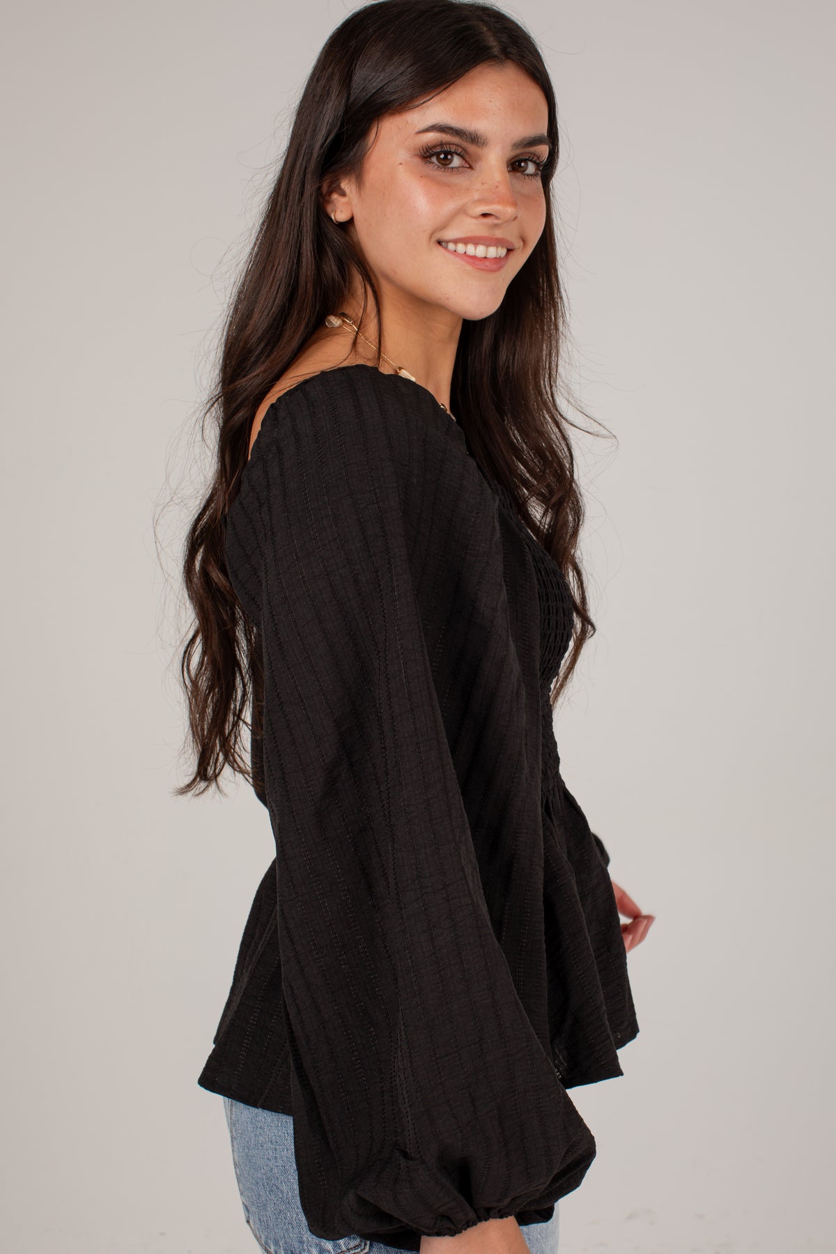 A woman wearing a black off-the-shoulder long-sleeve blouse with a textured crinkle fabric, paired with light blue jeans, photographed against a neutral light gray background.