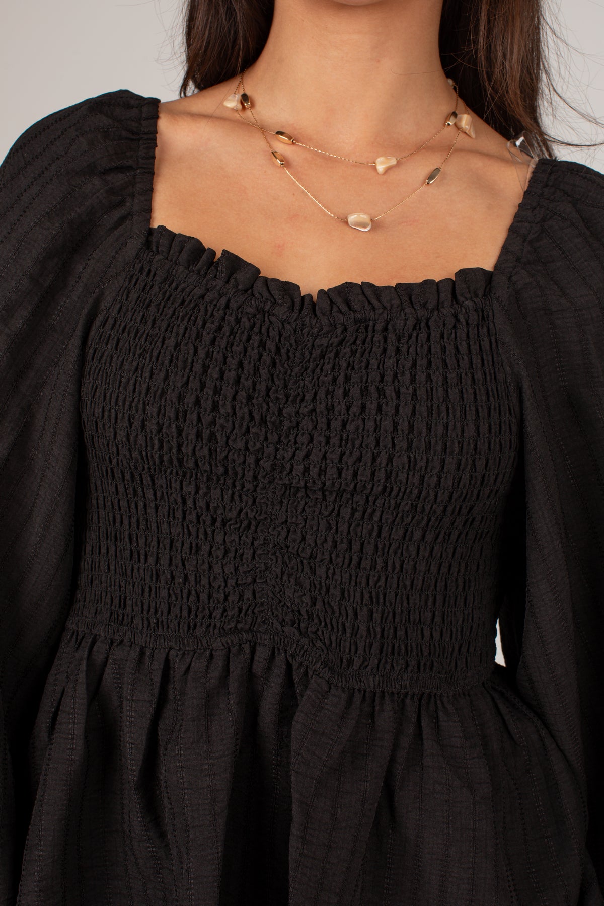 Close-up of a person wearing a black smocked long-sleeve dress with a square neckline and layered gold necklaces.