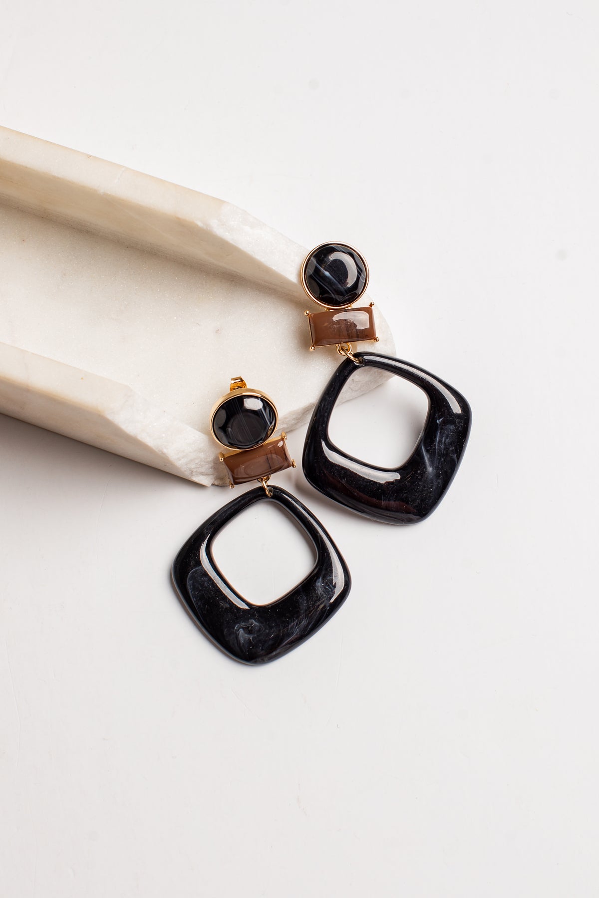A pair of statement drop earrings with large black marbled square dangles, gold-tone posts, and round black stone studs, displayed on a cream ceramic dish against a white background.