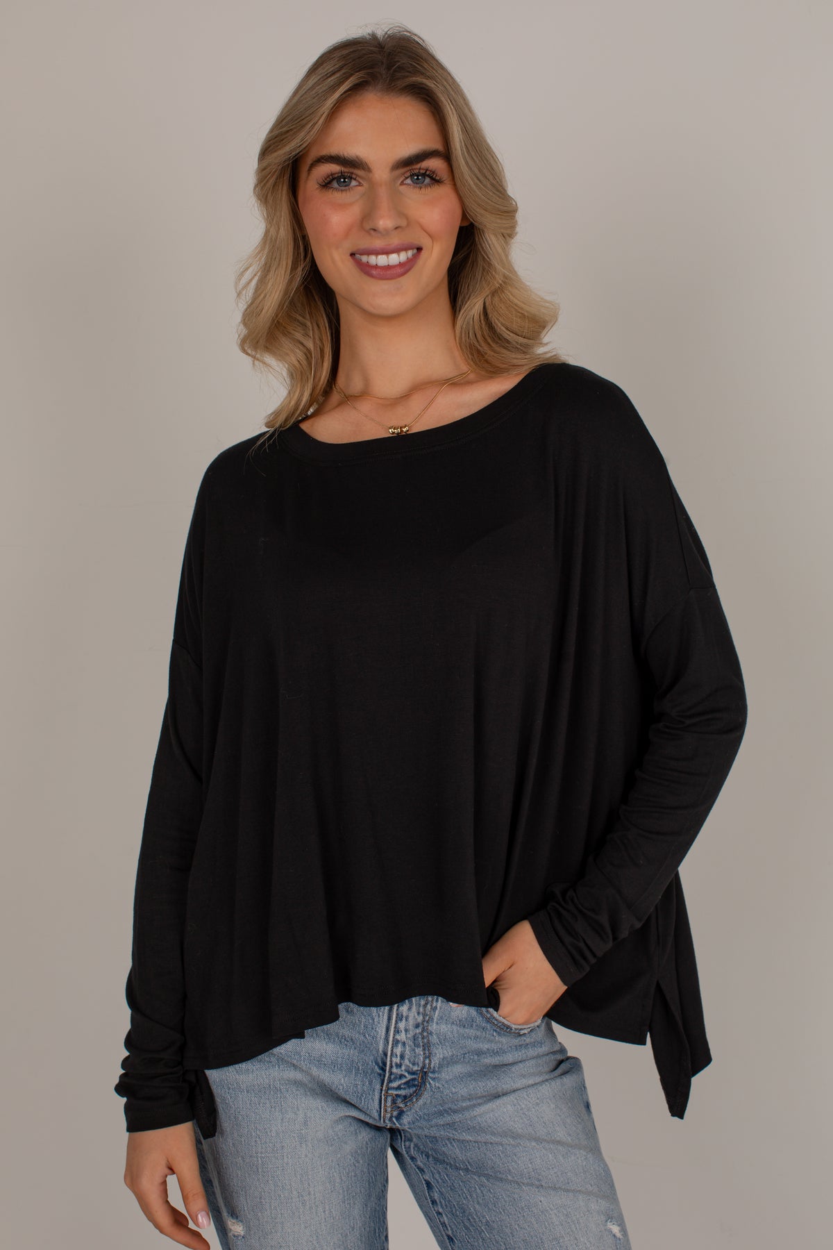 A woman with blonde hair wearing a black long-sleeve boat-neck top and light-wash blue jeans, standing against a solid light gray background.