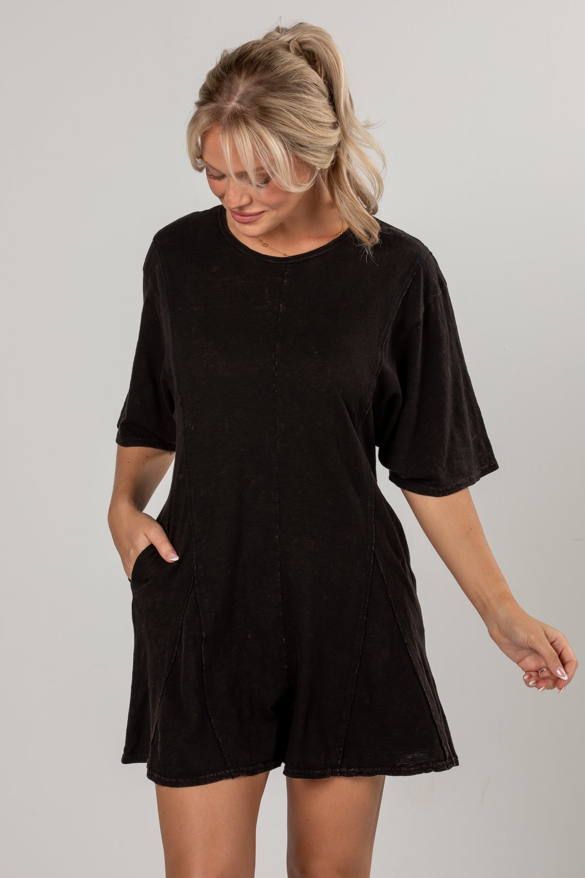 A woman wearing a black short-sleeve romper with side pockets, standing against a light gray studio background.