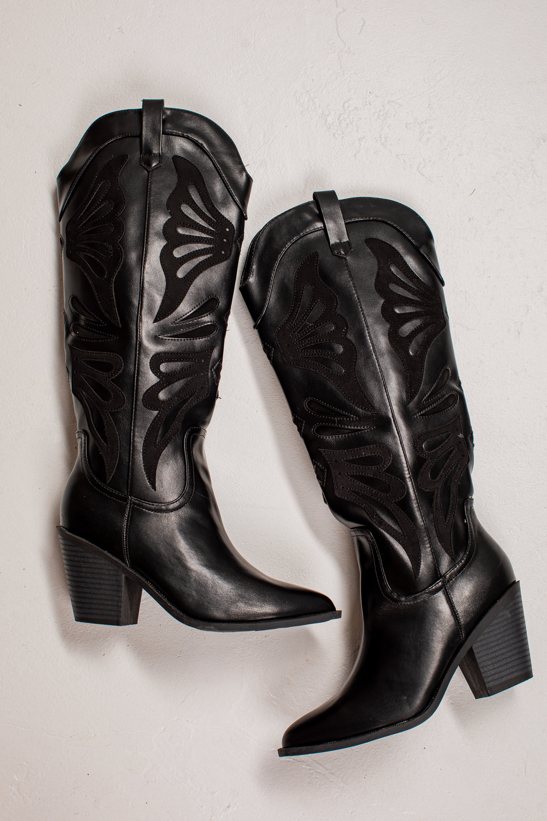 A pair of black leather knee-high cowboy boots with decorative cutout stitching, displayed on a light textured background.