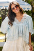 Woman wearing a light blue blouse with lace details and sunglasses, standing outdoors.