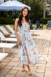 Woman in a floral dress standing on a patio with lounge chairs and an umbrella in the background.