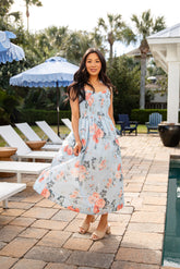 Woman in a floral dress standing by a poolside with lounge chairs and umbrellas in the background.