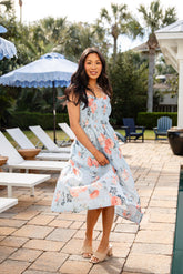 Woman in a floral dress standing by a poolside with lounge chairs and umbrellas.