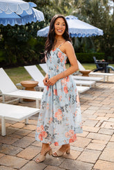 Woman in a floral dress standing in an outdoor setting with lounge chairs and umbrellas.