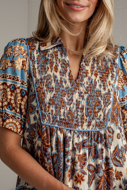 A close-up of a woman wearing a short-sleeve bohemian-style blouse with a smocked chest panel and a blue, brown, and cream floral pattern.