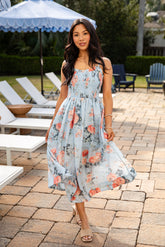 Woman in a floral dress standing outdoors near lounge chairs and an umbrella.