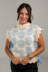 A smiling woman wearing a cream knit sweater vest with a light blue floral pattern, scalloped edges, and blue side ties, paired with black pants.