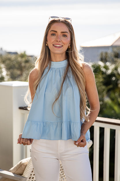 Woman wearing a light blue sleeveless top and white pants on a balcony with greenery in the background.