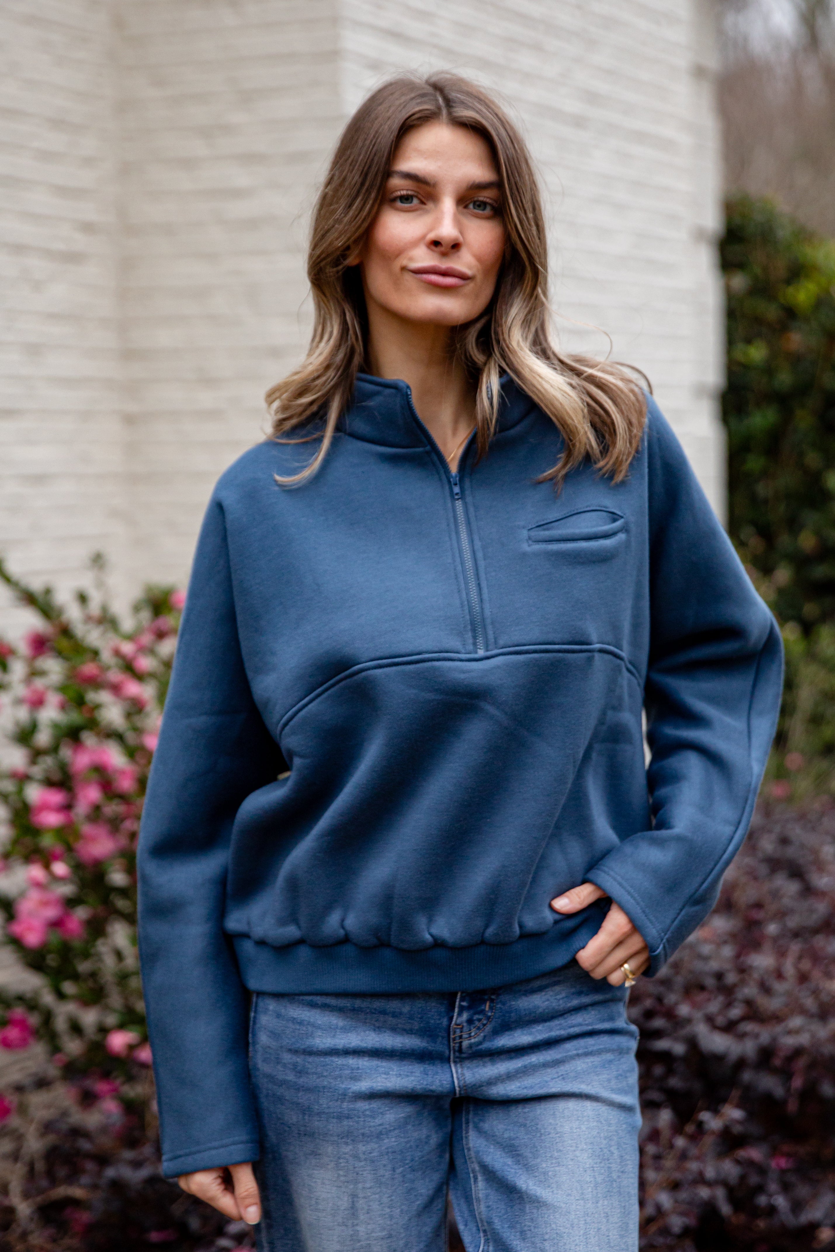 Woman wearing a blue pullover with a white wall and pink flowers in the background