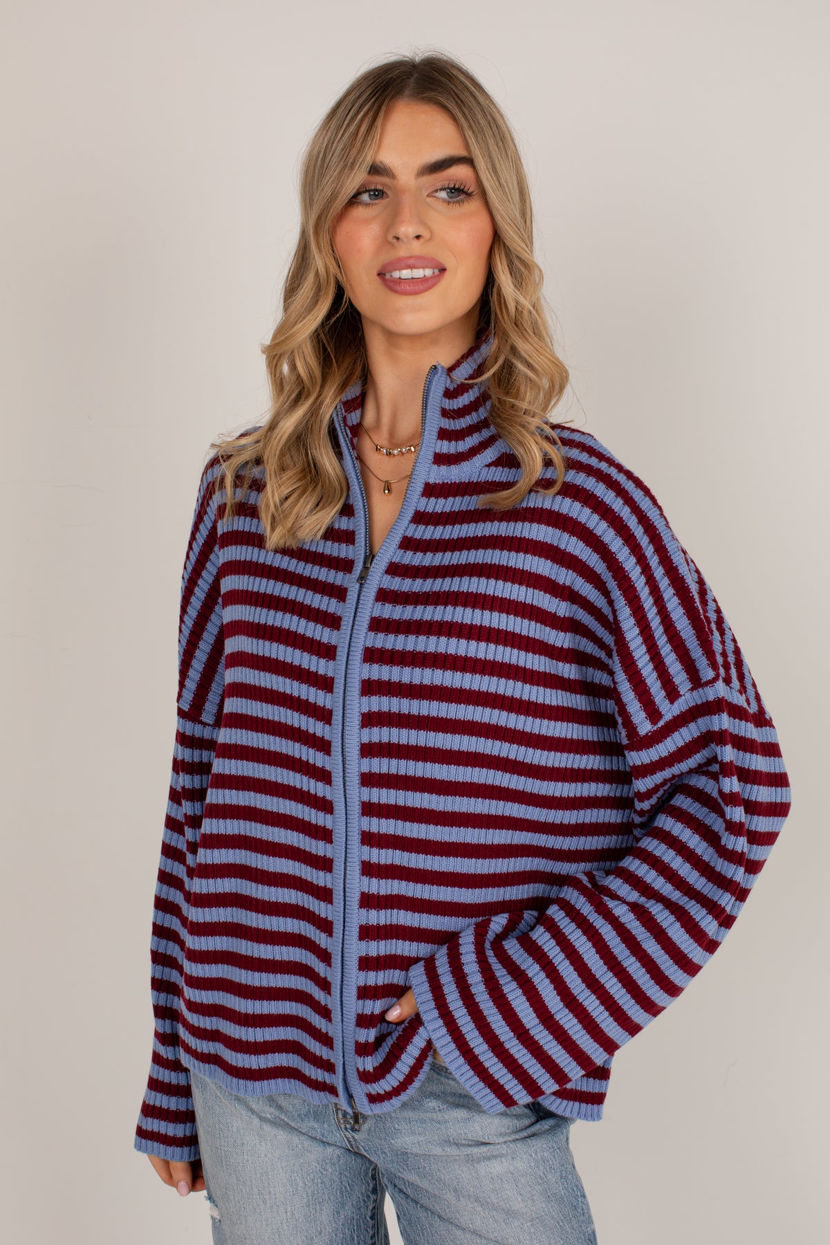 A woman modeling a waffle-knit zip-up sweater with horizontal stripes in burgundy and light blue, paired with light-wash jeans against a plain beige studio background.