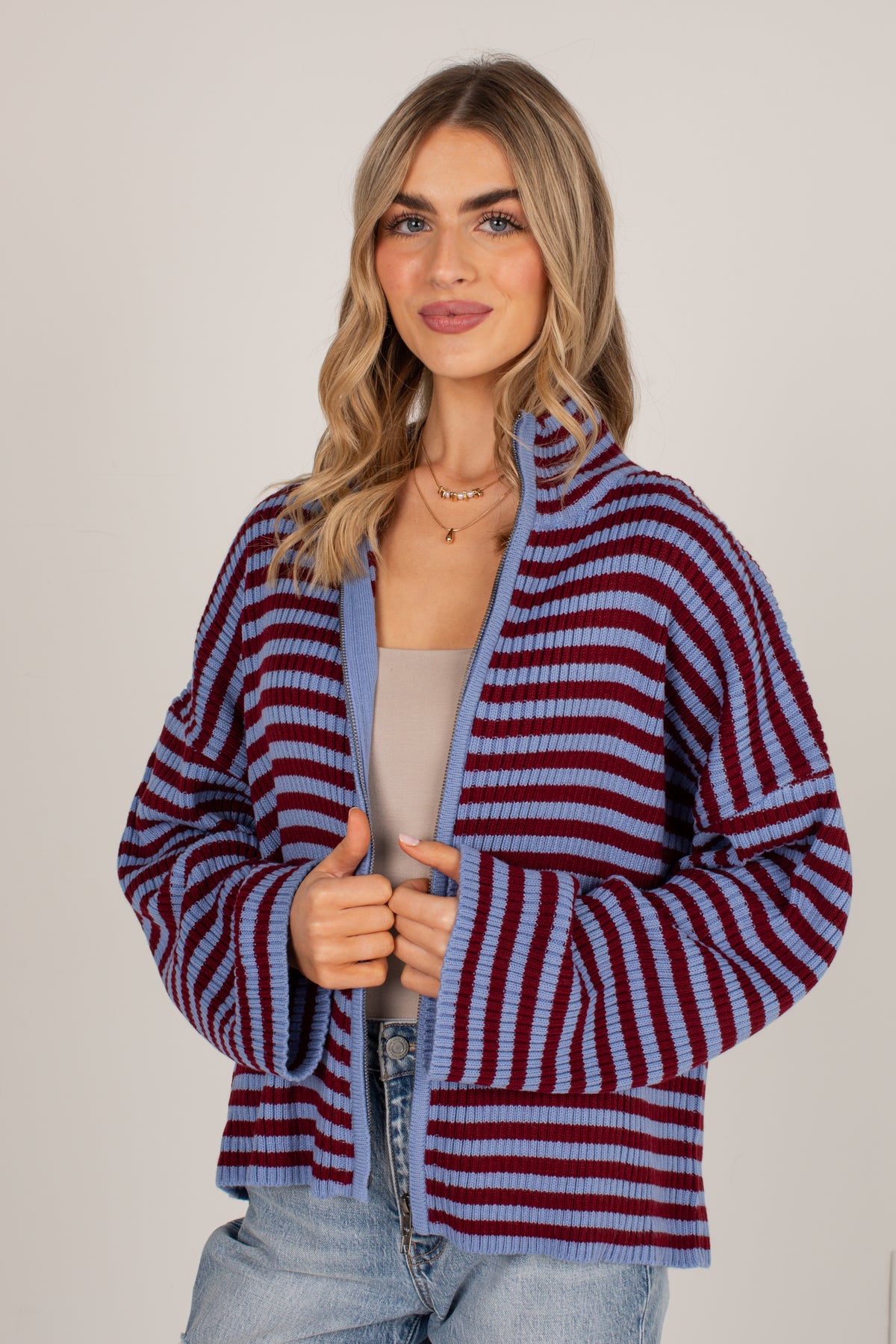 A woman modeling a striped knit cardigan with horizontal bands of burgundy and light blue, worn over a beige top and light wash jeans against a plain light gray studio background.