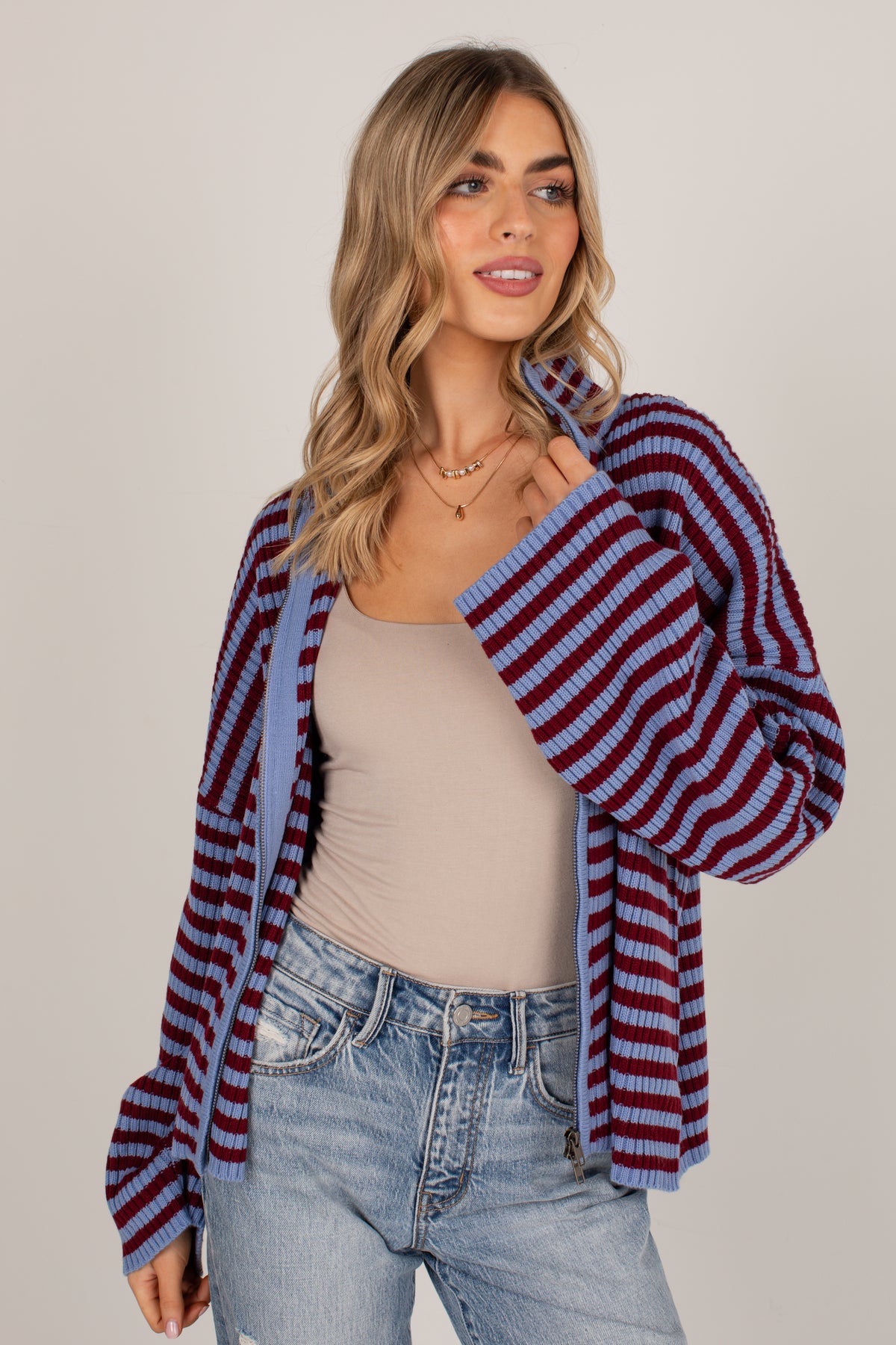 A woman modeling a striped knit cardigan with blue and burgundy horizontal stripes over a beige tank top and light blue jeans, set against a plain light gray studio background.
