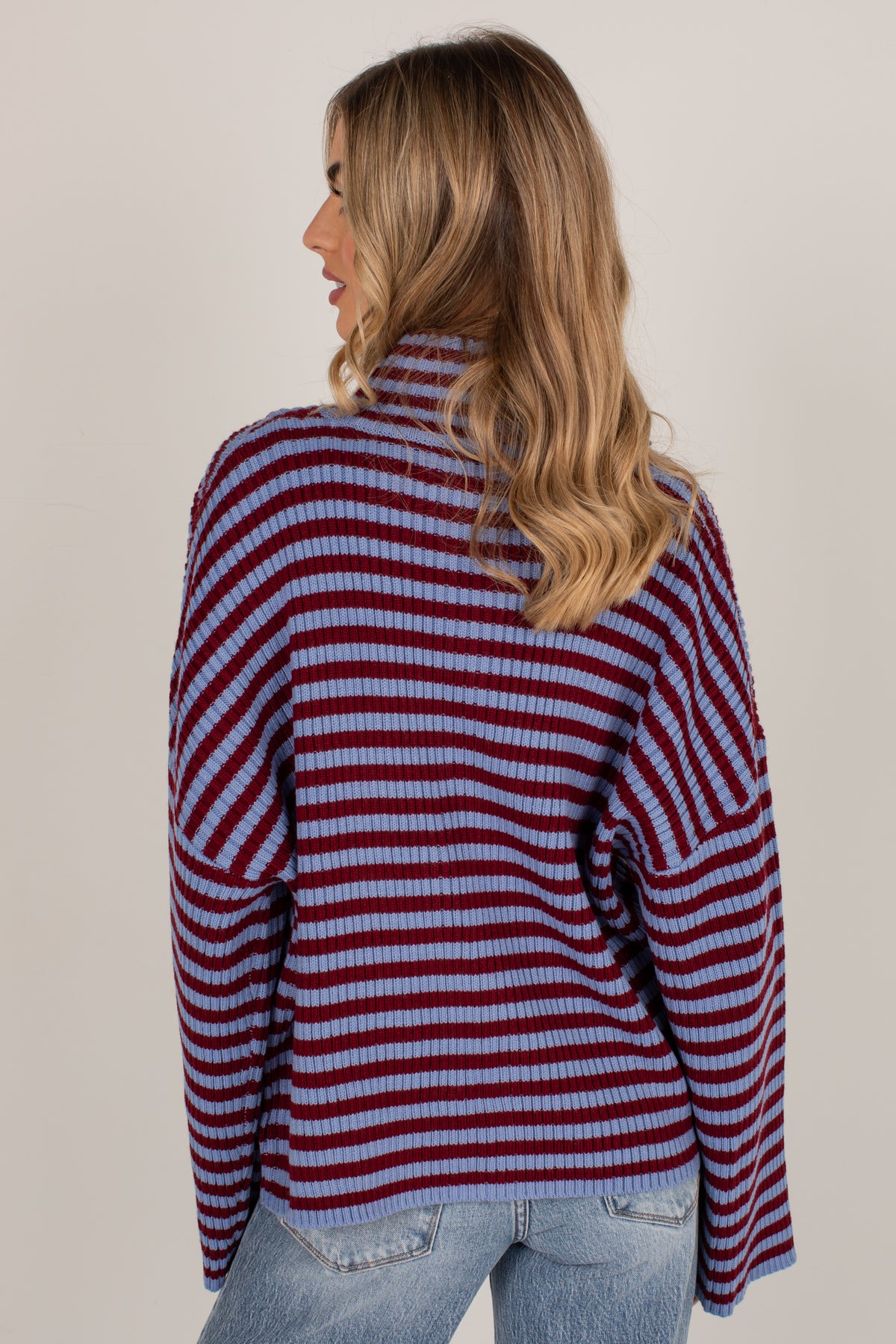 A woman with blonde hair is shown from the back wearing a ribbed knit mock neck sweater with horizontal burgundy and light blue stripes, paired with light wash blue jeans, against a plain light beige background.