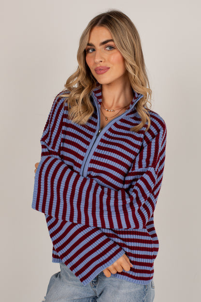 A woman wearing a long-sleeve quarter-zip knit sweater with horizontal burgundy and light blue stripes, paired with light-wash jeans, against a solid light gray studio background.