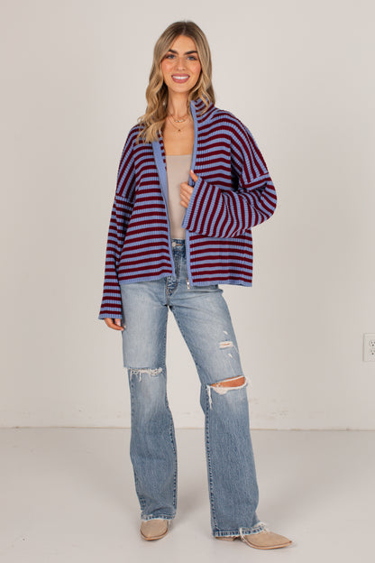 A woman modeling a striped cardigan with red and navy horizontal stripes, worn over a beige tank top and paired with light-wash distressed jeans and beige boots, against a plain off-white studio background.