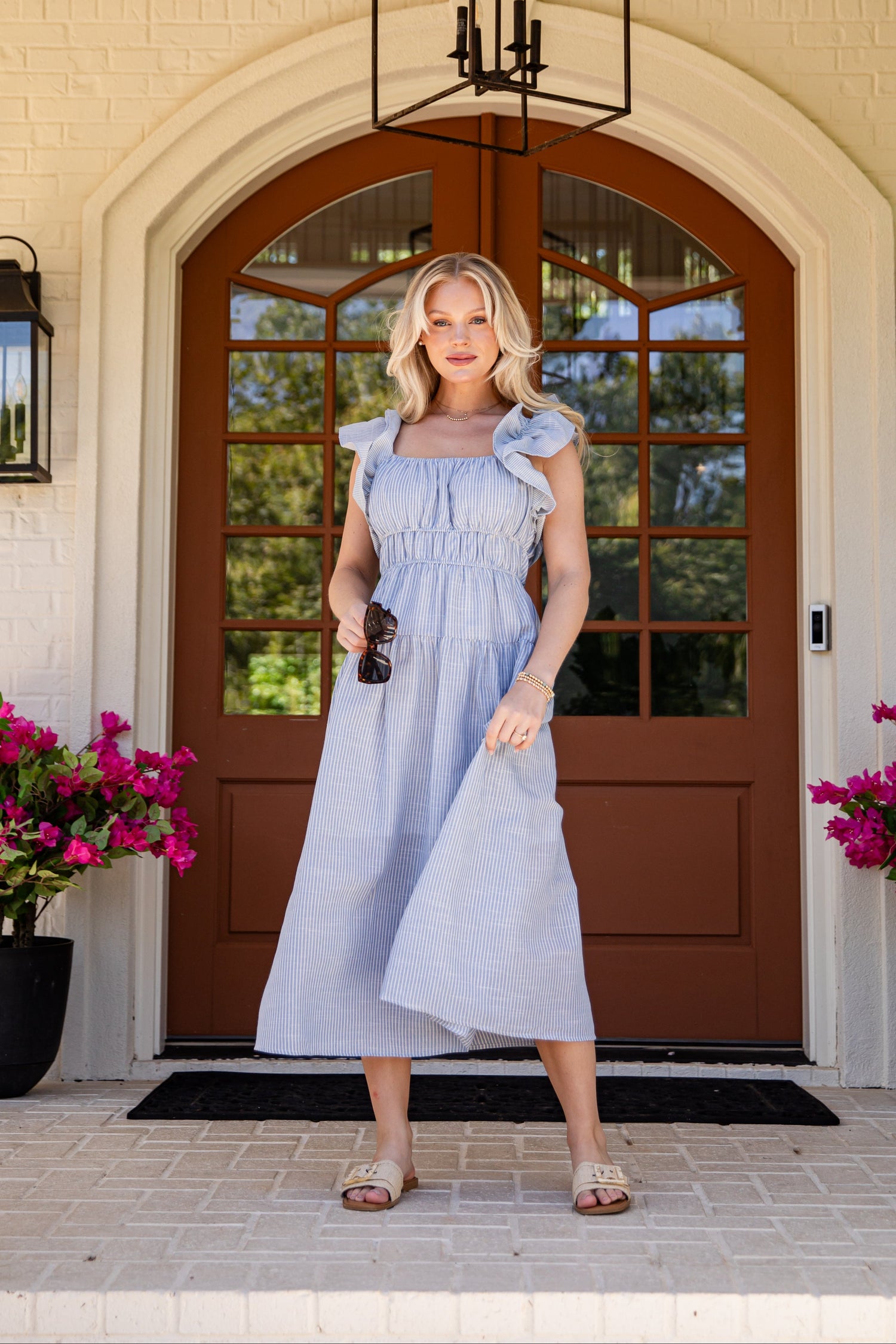 Woman in a light blue dress standing in front of a house entrance with pink flowers on either side.