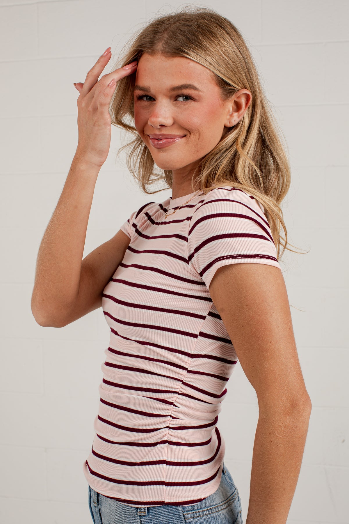 A woman wearing a fitted short-sleeve t-shirt with horizontal burgundy stripes on a light pink base, paired with blue jeans, posing against a white brick wall.