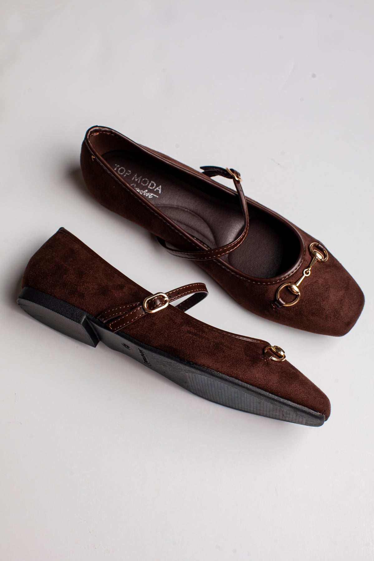 A pair of dark brown suede Mary Jane flats with gold-tone horsebit hardware and a low block heel, displayed on a light gray background.