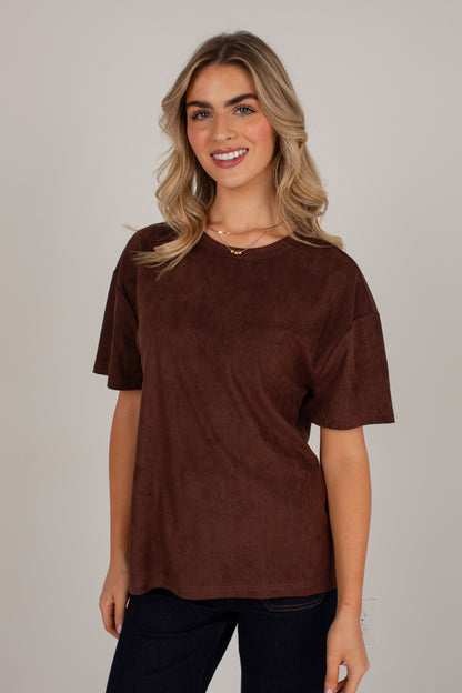 A woman wearing a brown short-sleeve crewneck top with a textured suede-like finish, paired with dark pants, against a solid light gray background.
