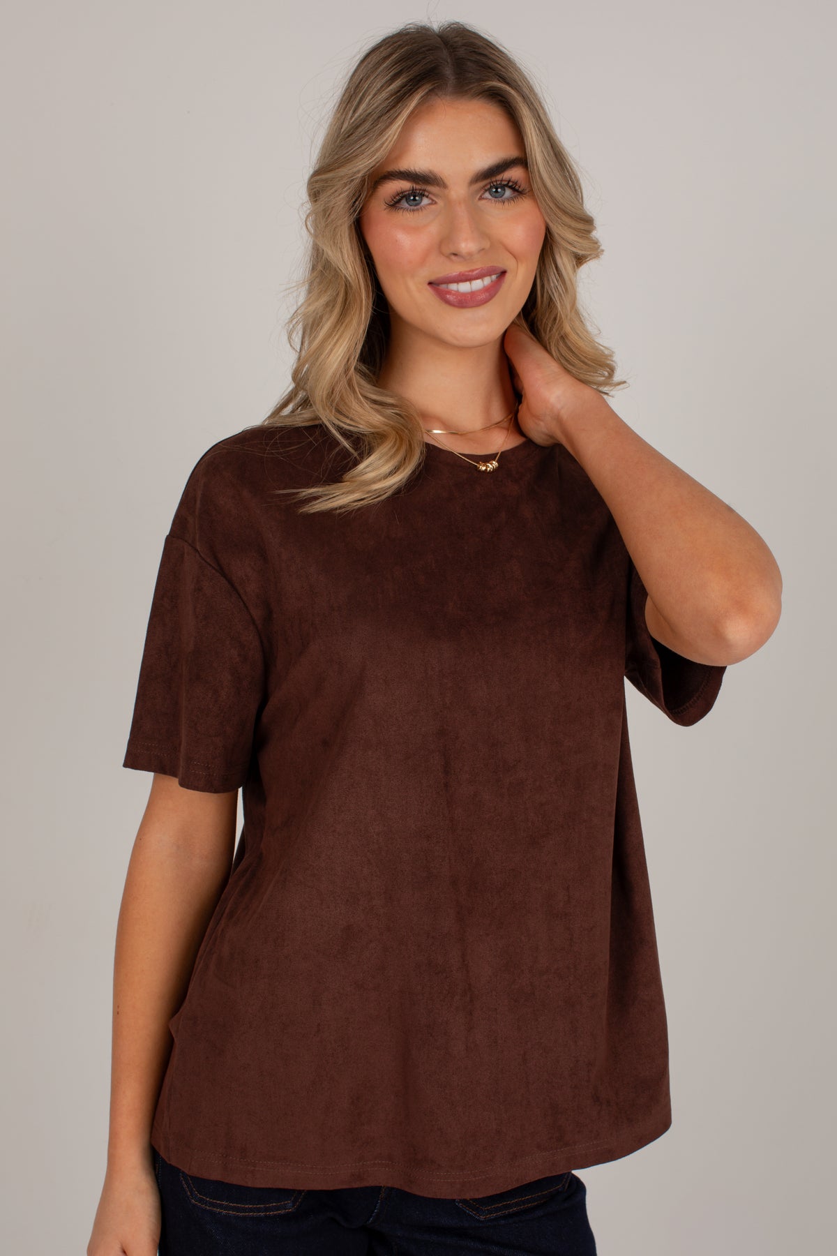 A woman wearing a brown short-sleeve top with a soft suede-like texture, posed against a solid light gray background.