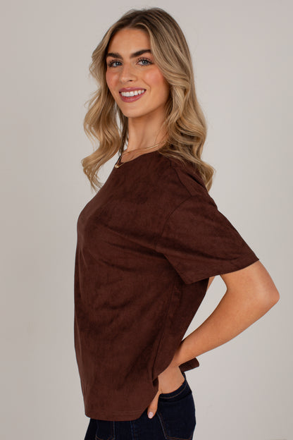 A woman modeling a brown short-sleeve top with a soft suede-like texture, paired with dark blue jeans, against a plain light gray studio background.