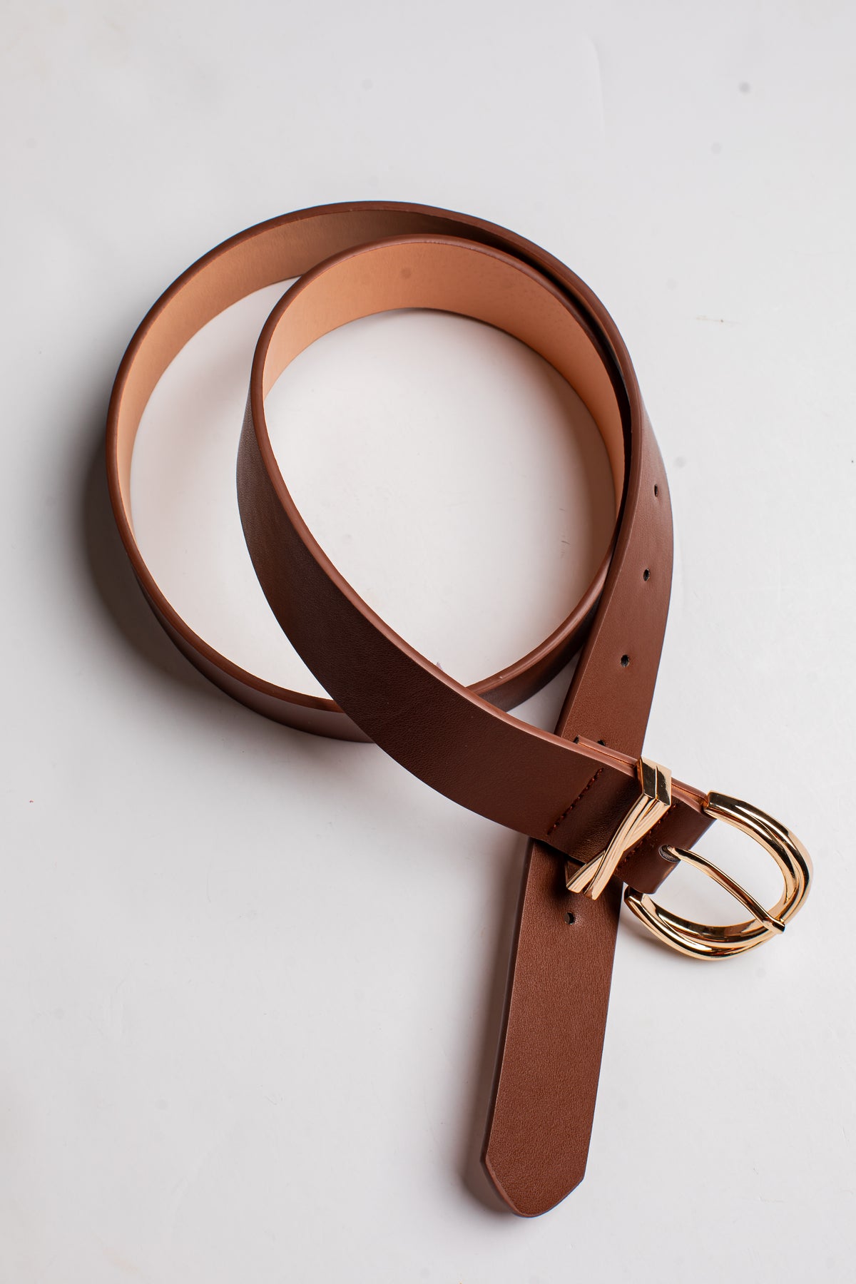 A brown leather belt with a gold-tone double-ring buckle, coiled on a light off-white background.