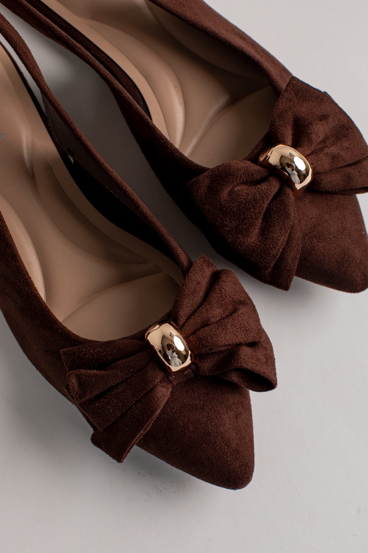 A pair of dark brown suede pointed-toe flats with decorative bows and shiny gold metal accents, shown on a neutral light gray background.