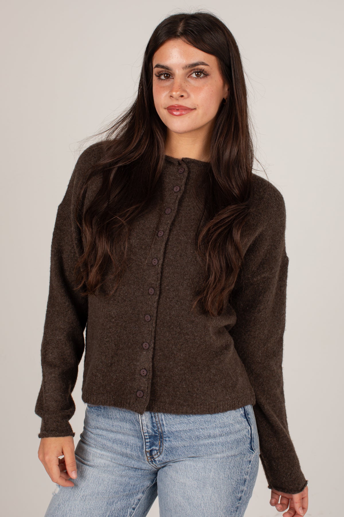 A woman with long dark brown hair wearing a dark brown button-up knit cardigan sweater and light wash blue jeans, standing against a plain light gray studio background.
