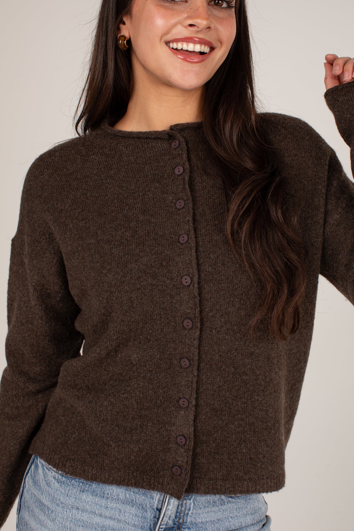 A woman wearing a dark brown button-up knit cardigan sweater and light blue jeans, smiling against a plain light beige background.