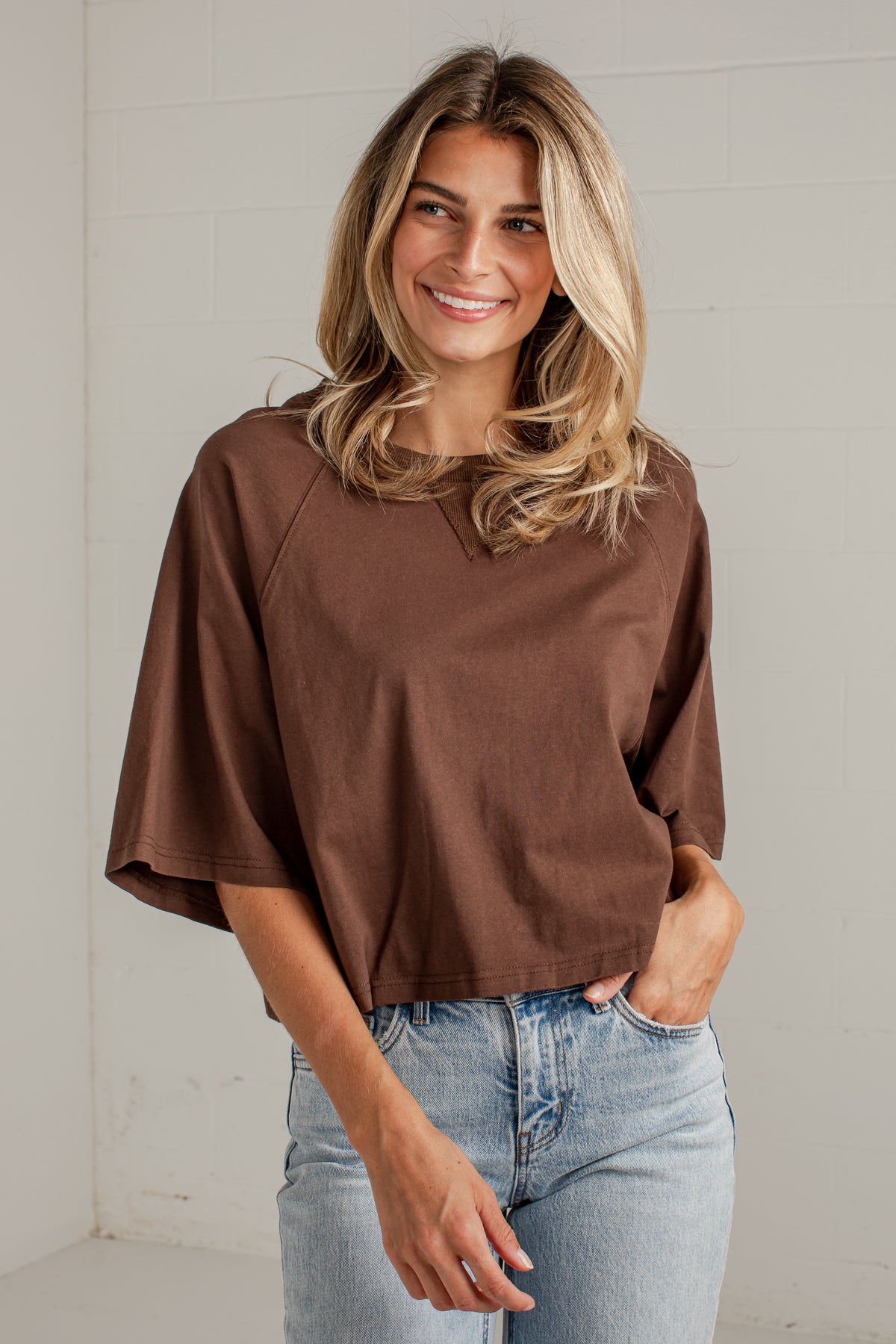 A woman wearing a brown short-sleeve top and light blue jeans, standing against a white brick wall.