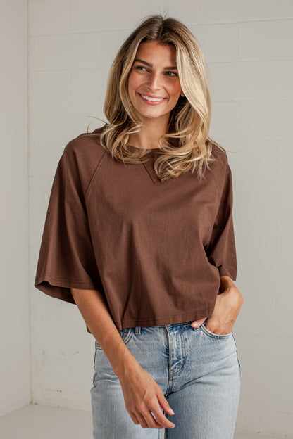 A woman wearing a brown short-sleeve top and light blue jeans, standing against a white brick wall.